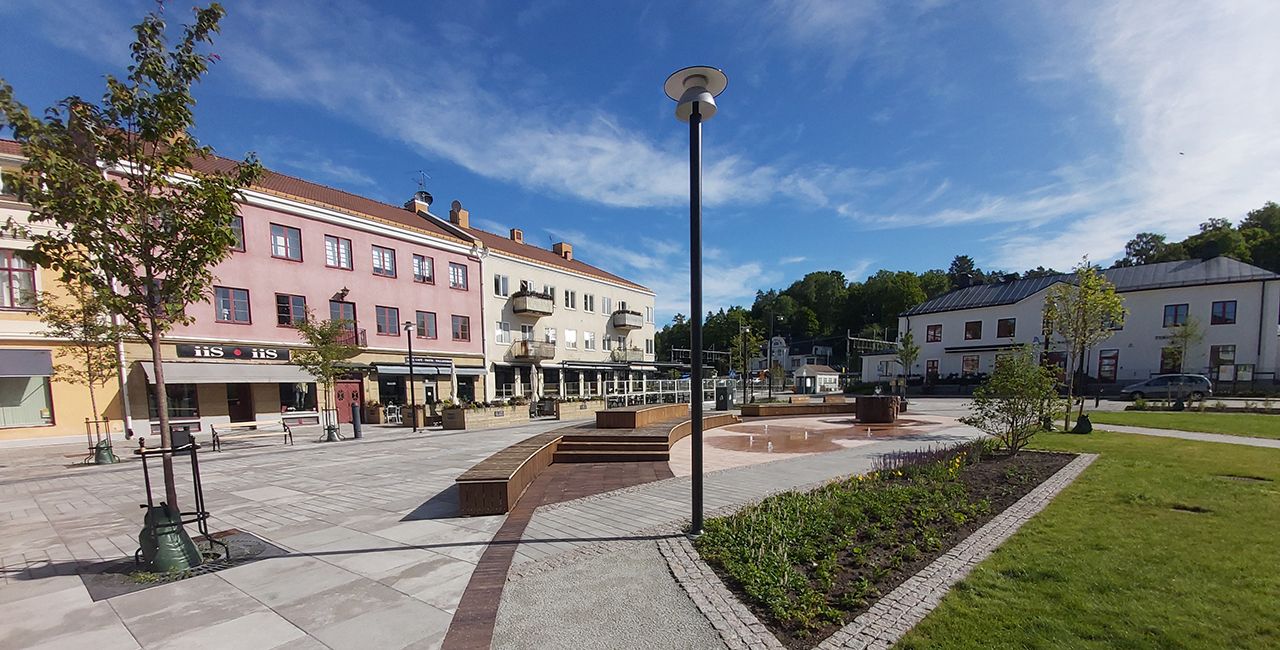 Ett torg med rosa och beige byggnader, grönytor och träd under en blå himmel.