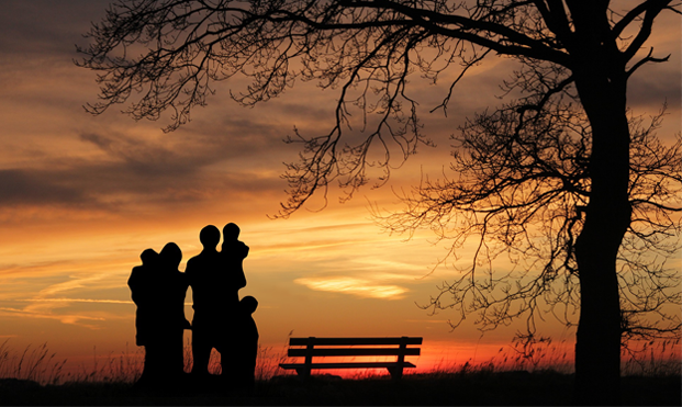 Två vuxna och tre barn syns som silhuetter mot solnedgången. De står under ett träd vid en parkbänk.