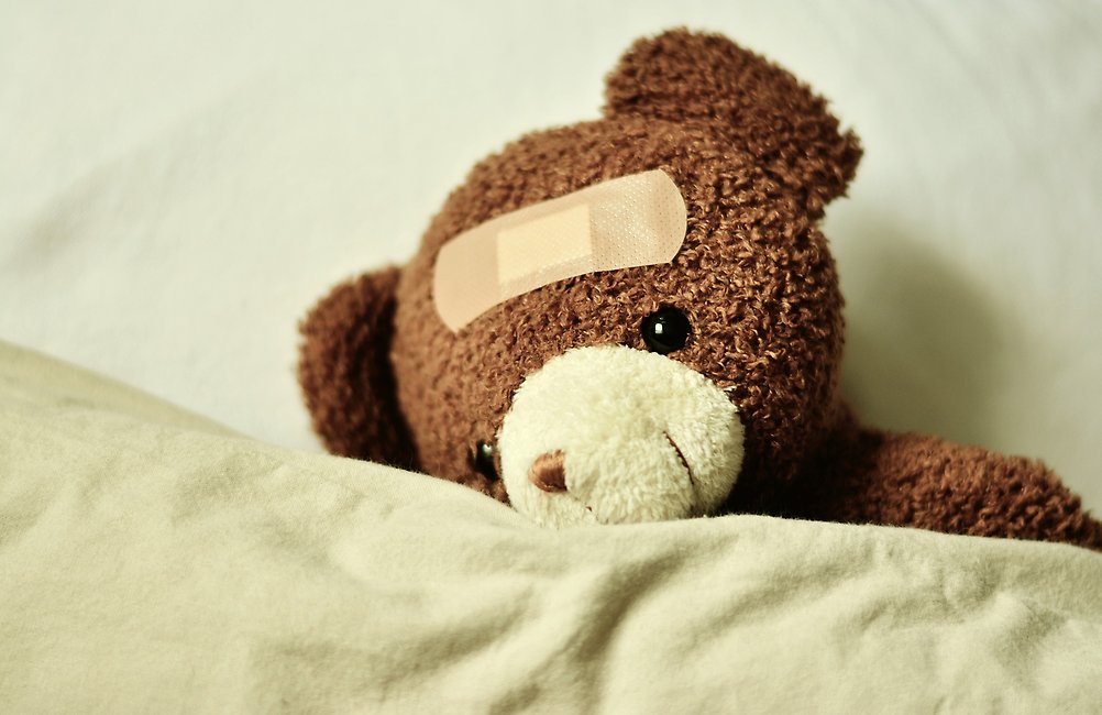 En nallebjörn som ligger under ett täcke och som har ett plåster i pannan.